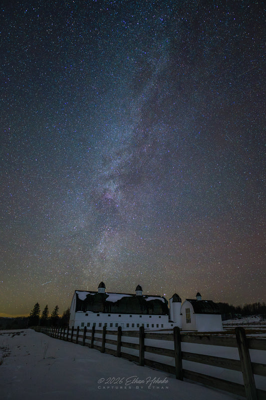 Milky Way galaxy over a snowy landscape with a white building and wooden fence.